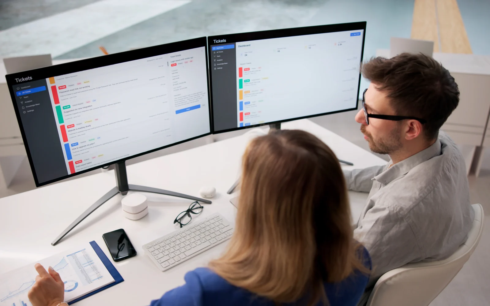 Two colleagues reviewing a service desk queue on dual monitors